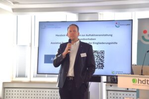 Marco Schmitz hält eine Rede. Hinter ihm ist der Ausschnitt einer Präsentation zu sehen.